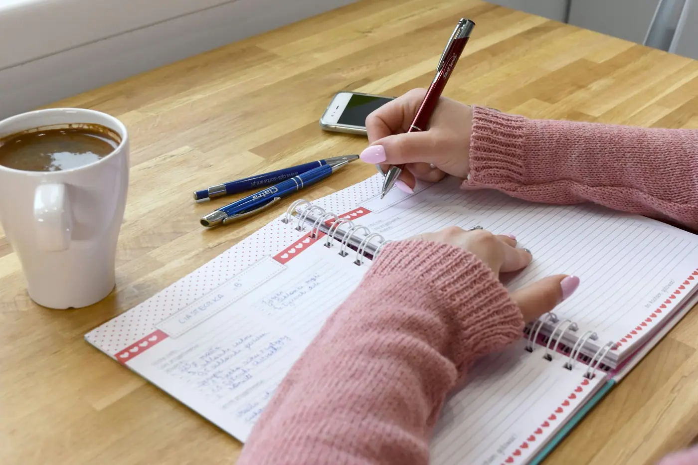Une personne écrit dans un carnet sur une table en bois, une tasse de café à côté